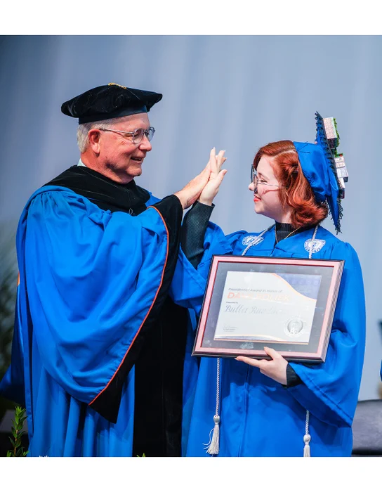 North Carolina State Auditor Dave Boliek gives Randolph Community College graduate Bullet Rundberg a high five during the 2026 Commencement Ceremony. Rundberg, dressed in blue graduation regalia and holding a framed award presented in honor of Boliek, smiles on stage beside RCC President/CEO Dr. Shah Ardalan.