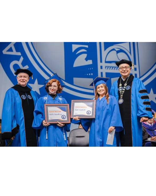 Graduates Bullet Rundberg and Ruth Lambeth stand on stage with framed awards presented in honor of North Carolina State Auditor Dave Boliek during Randolph Community College’s 2026 Commencement Ceremony. RCC President/CEO Dr. Shah Ardalan stands between them while speaking with the graduates.