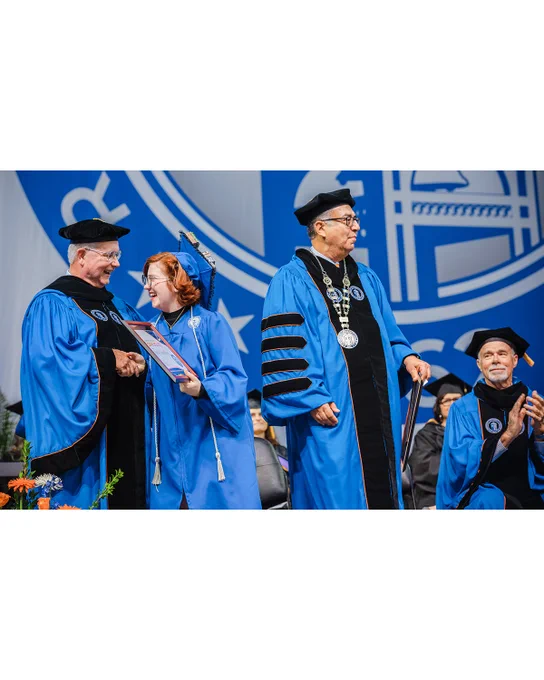 Randolph Community College graduate Bullet Rundberg smiles while receiving a framed $1,000 award presented in honor of North Carolina State Auditor Dave Boliek during RCC’s 2026 Commencement Ceremony. RCC President/CEO Dr. Shah Ardalan stands beside them on stage.