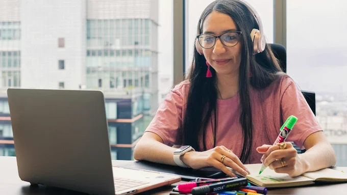 A person is watching a laptop screen and wearing headphones. They are highlighting something in a notebook.