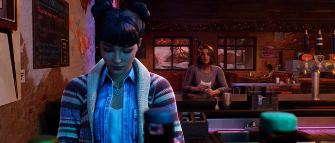 Amanda Thomas from Life is Strange: Double Exposure, an Indigenous woman of Cherokee and Algonquin descent, standing behind the counter inside her bar, The Snapping Turtle. She is looking down with a focused and serene expression. Her dark hair is styled in space buns with blunt bangs, and she wears a denim shirt under a striped knit cardigan. A soft, cool blue light from a nearby aquarium illuminates the side of her face, contrasting with the warm, dimly lit interior of the Lakeport bar. In the background, Max Caulfield sits at the bar counter, watching Amanda attentively. Further back, Professor Gwen Hunter is visible sitting alone at a dimly lit table near the window.