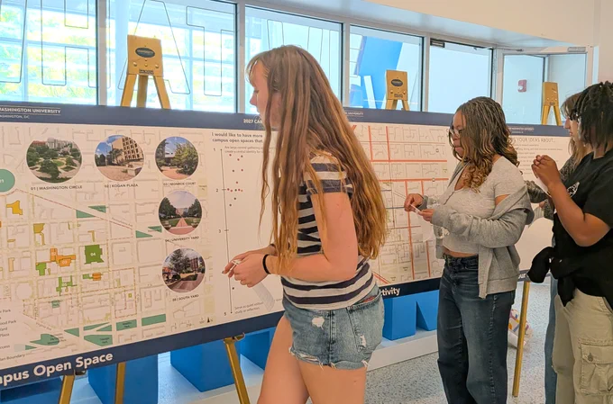 Three people are viewing and interacting with detailed maps and charts on easels related to Washington University. The background includes large windows displaying the university's name in part. The scene takes place indoors, suggesting an academic or planning event.