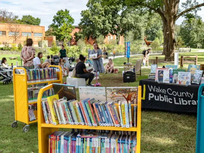 Outdoor community event with Wake County Public Library bookshelves and people gathered on grass near trees.