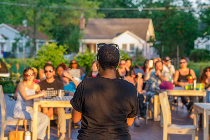 Person speaking to an attentive outdoor audience seated at tables in a sunny garden setting.