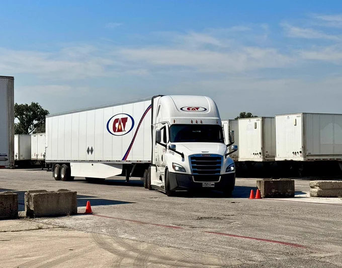 White C.A.T. Inc semi truck with a long trailer featuring the red and blue CAT logo drives through a yard lined with parked white trailers. Orange safety cones and concrete barriers sit in the foreground on a paved lot under a bright blue sky with light clouds.