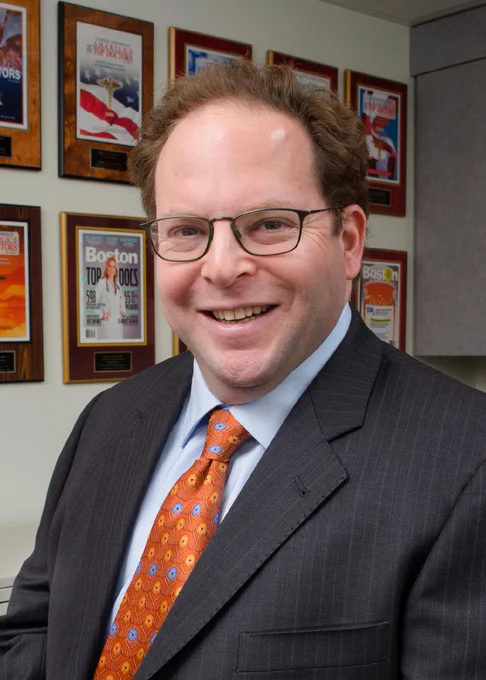 Man with glasses and an orange tie with a blue shirt and black blazer smiling to the camera. behind him are several plaques on the wall.