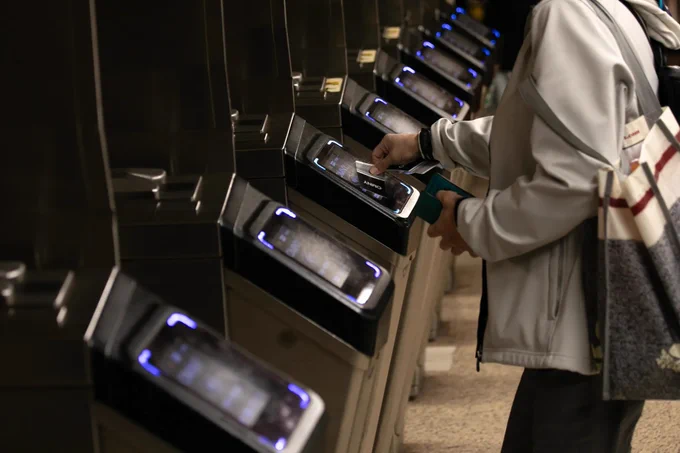 A person using a fare payment machine at a transit station turnstile, with several similar machines visible in a row. The person is wearing a light-colored jacket and carrying a bag.