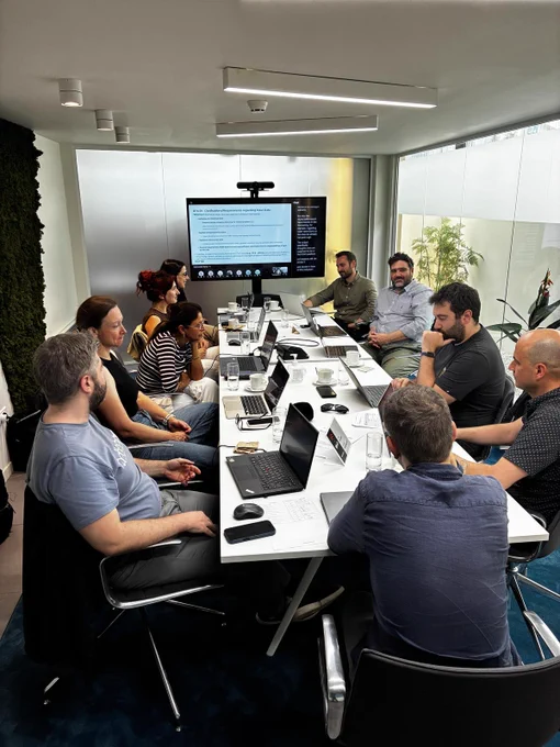 Image shows a group of people sitting at a table with laptops. A screen is at the top of the table.