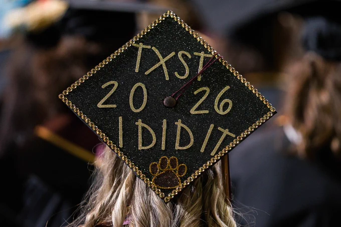 Back of a graduation cap decorated in gold glitter letters with text reading "TXST 2026 I did it"