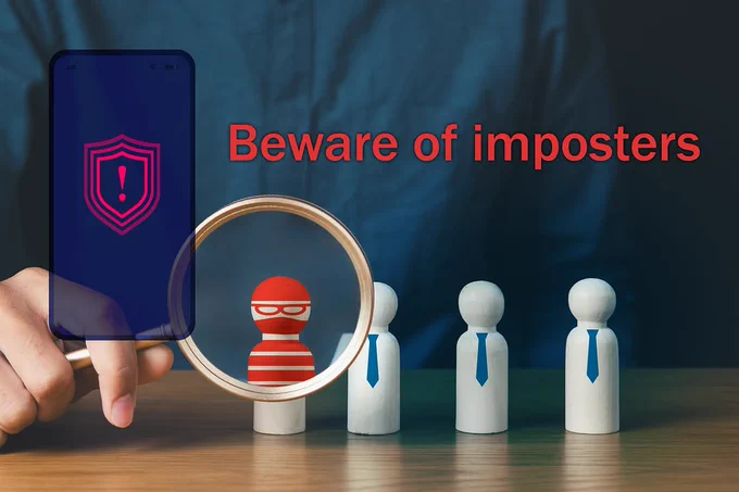 A person holds a magnifying glass over a group of wooden figures, revealing a red masked figure among others wearing neckties. A smartphone screen shows a red shield with an exclamation mark. The text "Beware of imposters" is prominently displayed.