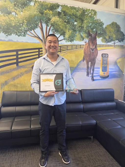 A person holds a certificate and award in front of a scenic mural depicting a horse and a robot on a path. The scene is cheerful and celebratory.