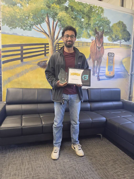 A person stands holding an award and certificate in front of a mural depicting a pastoral scene with a horse. The scene is calm and celebratory.