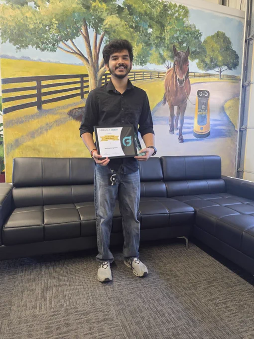 A person smiling and holding an award certificate. They are standing indoors in front of a mural featuring a horse and a robot.