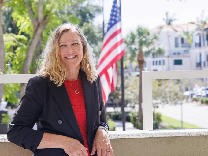 Supervisor of Elections Melissa R. Blazier posing for a picture in a black blazer and red top standing outdoors with an American flag and residential buildings in the background.
