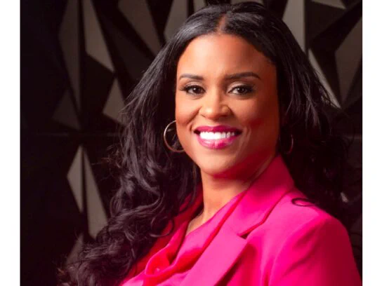 Woman with long wavy black hair wearing a bright pink blazer against a dark geometric background.