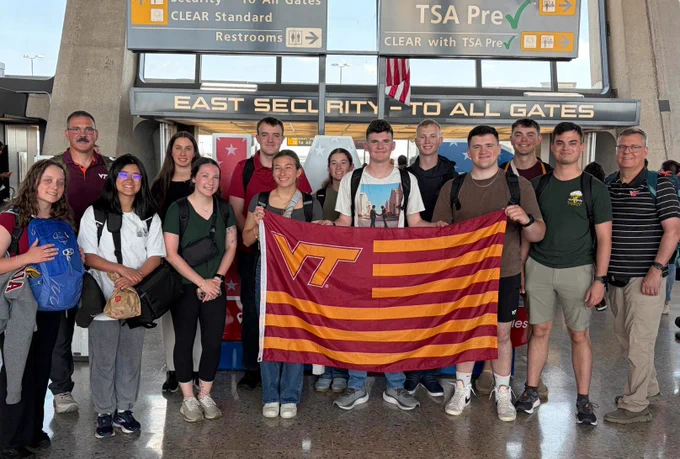 A large group of cadets and two staff members hold a striped Virginia Tech flag at an airport with TSA precheck signs in the background.
