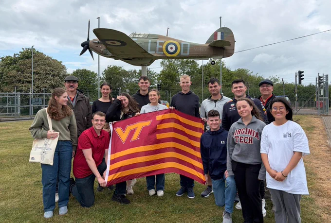 A large group of cadets and two staff members hold a striped Virginia Tech flag while standing in front of a static display of a WWII fighter plane