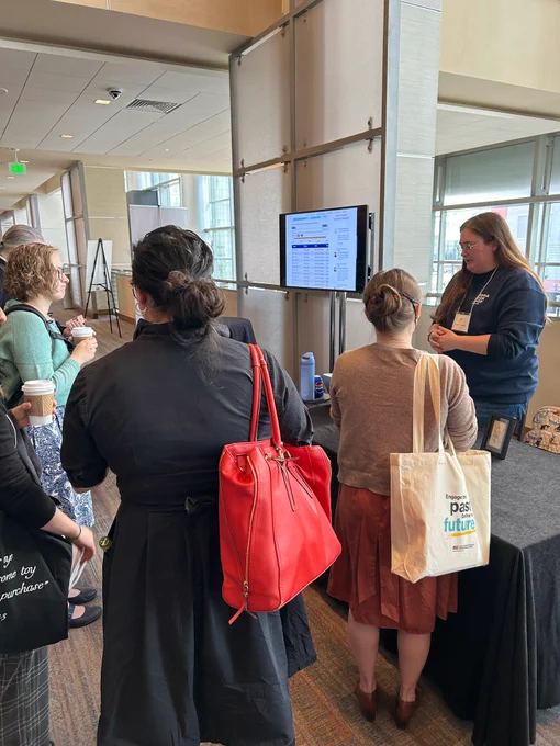 Image of several academics clustered around a table and computer monitor, listening to an early career academic introduce their digital project. End ID.