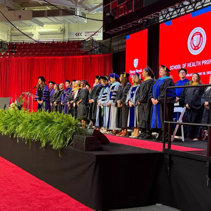 A group of people in academic regalia stand on a stage during a ceremony at Stony Brook University School of Health Professions. The stage is decorated with green plants, and a red backdrop displays the school's logo. Audience members are visible in the background.