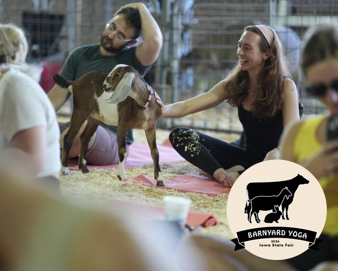 A group of people sitting on yoga mats smiling while sitting down and petting a goat. 