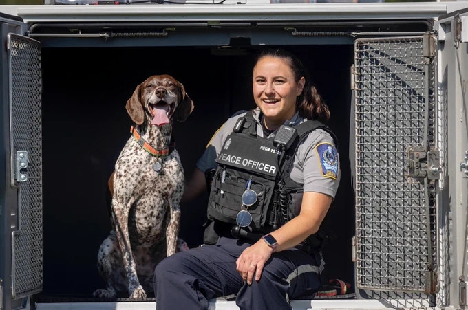 A smiling peace officer sits in the back of a vehicle transport kennel next to a happy German Shorthaired Pointer.