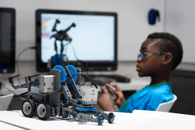 Close-up of a detailed robotic vehicle model on a desk with a child in the background using a controller and a computer screen showing the robot's image.