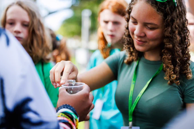 Person wearing a green shirt pouring a liquid into a clear glass.