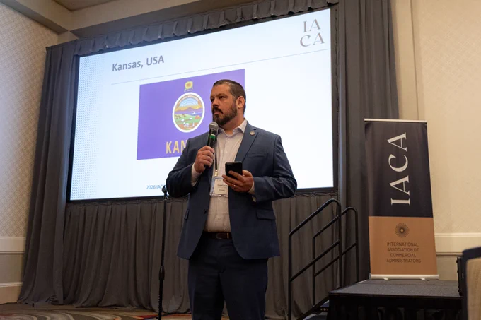 Man speaking at a microphone with screen behind him, with "Kansas" title and flag