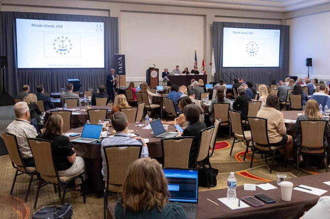 Large conference room with round tables at the front and more tables in the back, a stage and two large screens at the front 