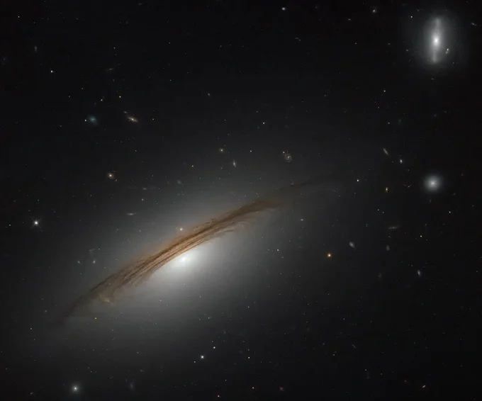 A massive spiral galaxy is seen slightly angled away from edge-on, as if we are viewing the underside of the disk. Dark dust lanes outline the shape while the bright bulge glows from the center. The galaxy is tilted from the lower right to the upper left and occupies two-thirds of the image. In the upper right, a much smaller and/or more distant barred spiral galaxy can be seen.