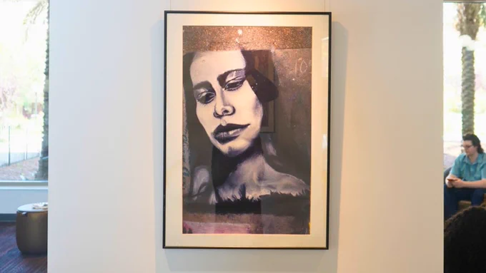 A framed artwork hangs on a wall, featuring a stylized portrait with strong contrasts in black, white, and purple tones. In the background, two people are seated on a bench.