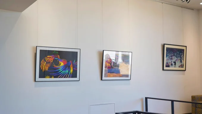 A white wall displays three framed artworks. The left artwork features a vibrant parrot with colorful feathers. The center piece shows an abstract scene with warm tones. The right artwork depicts a busy crowd in a city setting.
