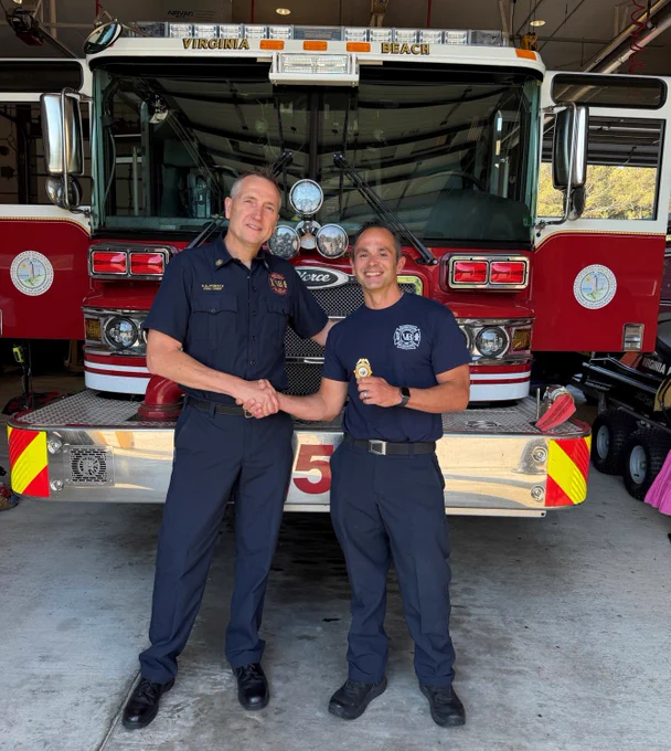 Two individuals in navy uniforms stand in front of a Virginia Beach fire truck. One person holds a badge and they are shaking hands in front of the vehicle's open garage.