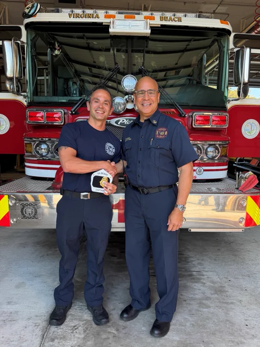 Two smiling people in uniform stand in front of a Virginia Beach fire truck. One person holds a fire helmet.