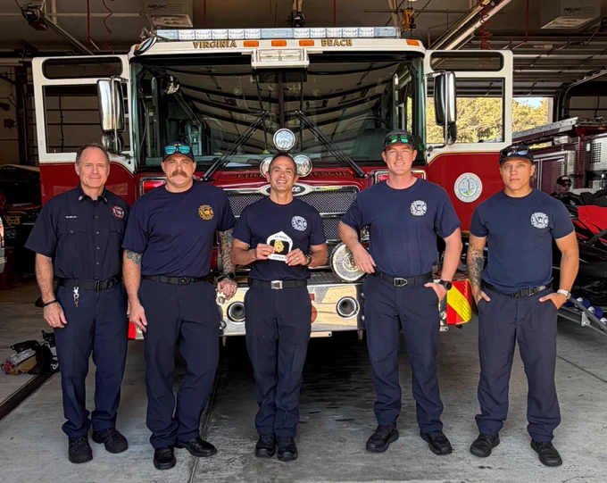 A group of five people in uniform stand in front of a Virginia Beach fire truck in a garage. They are smiling and one person is holding a small award.