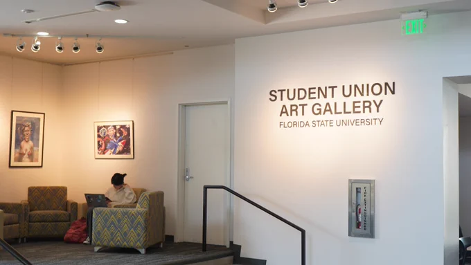 A person is seated on a cushioned chair in a lobby, using a laptop. The walls feature artwork, and signage reads "Student Union Art Gallery, Florida State University.