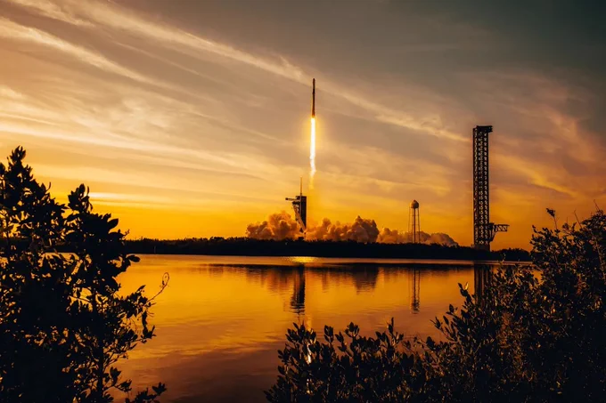 A rocket launches into the sky at sunset from Kennedy Space Center, reflected in calm water below. Dark silhouettes of trees frame the foreground while orange and gold clouds fill the sky.