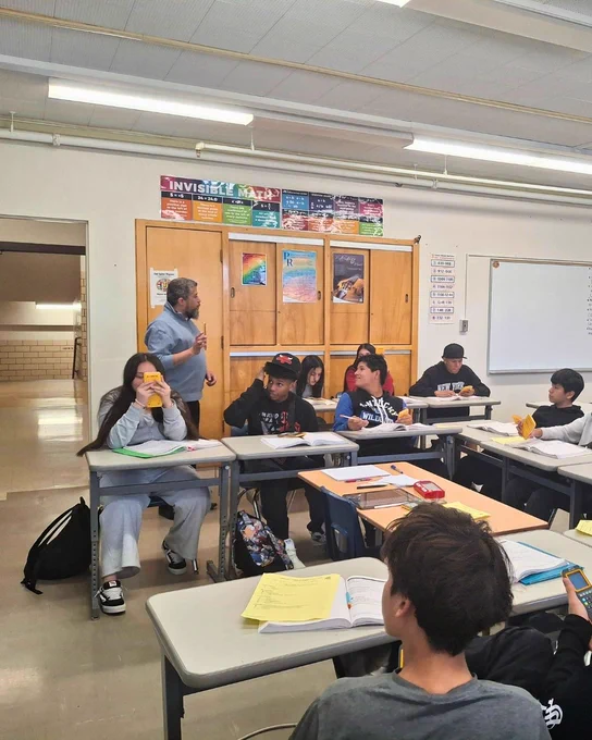 A classroom scene with several people sitting at desks, reading and writing. A person stands at the front, engaging with the group. Educational posters, including one featuring "Invisible Man," decorate the wall. There are open notebooks and highlighters on the desks.