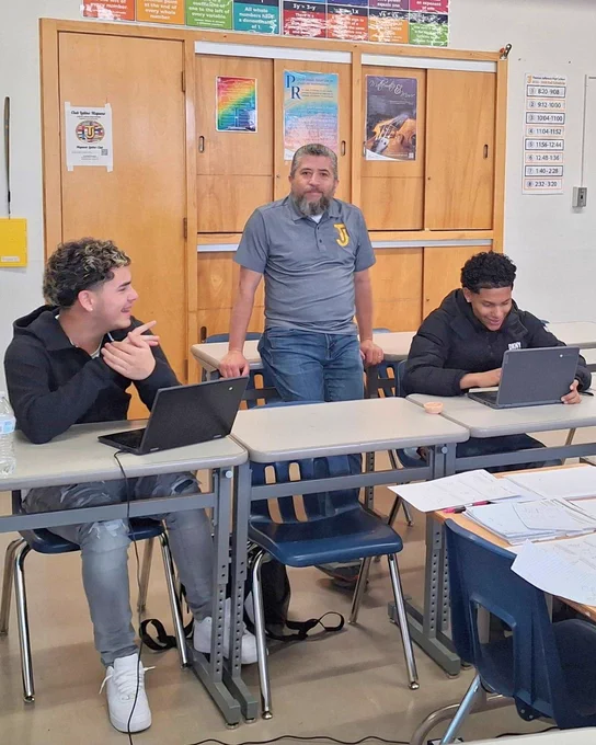 A person stands between two students seated at classroom desks with laptops. Colorful posters, including a motivational one, hang above wooden cabinets in the background. Papers and books are on the desks.