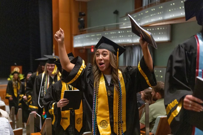A graduate in a black cap and gown adorned with yellow accents and honor cords enthusiastically raises her arms in celebration. She holds a diploma in one hand and appears joyful amid a procession of graduates in an auditorium setting.