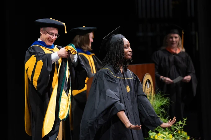 A graduate in a black cap and gown walks on stage with outstretched arms, smiling. A faculty member dressed in a black and gold academic robe prepares to place a hood around their shoulders. Other faculty members in similar attire stand in the background.
