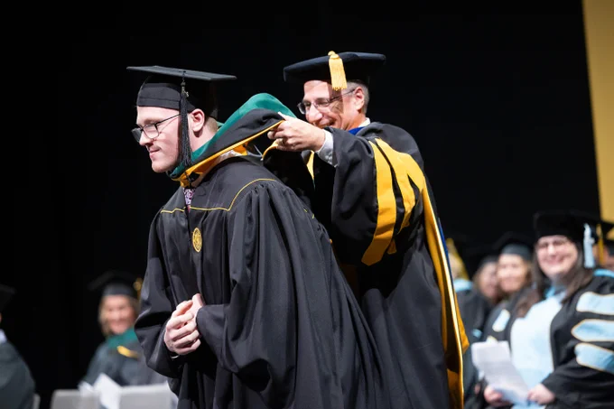 A graduate is being hooded by a faculty member during a commencement ceremony. The graduate wears a black cap and gown with a medallion, while the faculty member, in academic regalia with yellow accents, places a green and gold hood over the graduate's shoulders. Other faculty members, also in caps and gowns, are visible in the background, smiling and watching the ceremony.