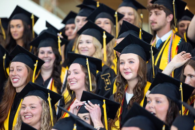 A group of graduates wearing black caps and gowns with yellow sashes stands together, smiling broadly. The image captures a joyful moment, with tassels hanging from their caps and a diverse group of faces all focused in the same direction.