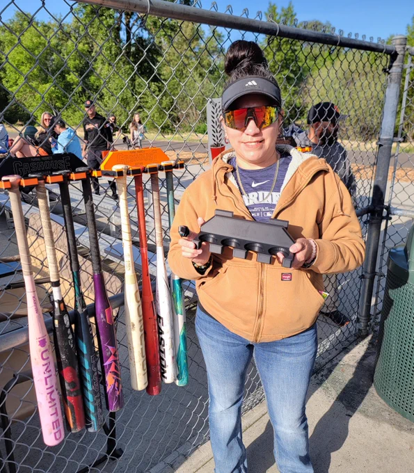 Adriana, an RCC student, stands proudly at her daughter's baseball game with her handmade baseball bat holder in hand. She wears a khaki jacket, jeans, sunglasses and visor.
