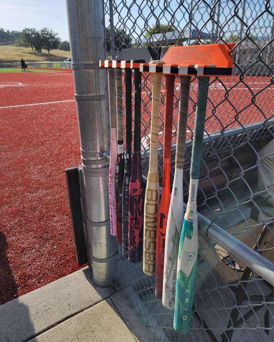 A collection of baseball bats hanging vertically on a fence near a baseball field with red artificial turf. The bats feature various designs and brands, and sit in a custom holder manufactured by an RCC student. The field is surrounded by fencing and grass can be seen in the distance.