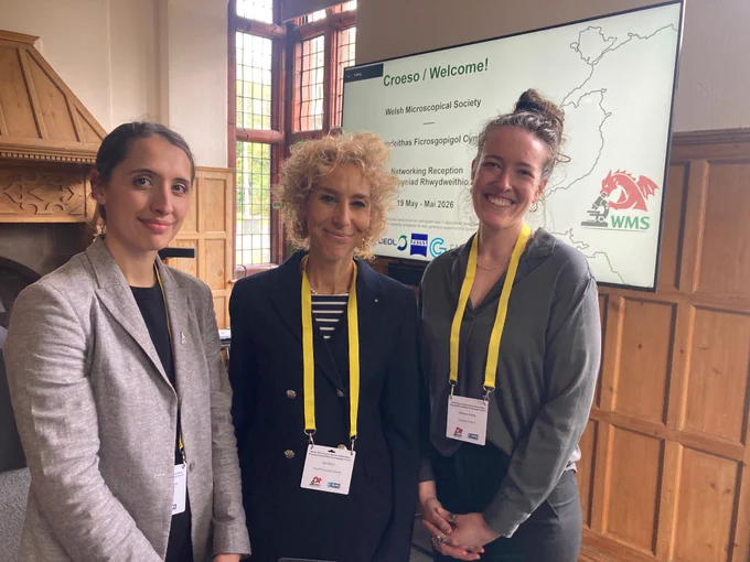 Three attendees wearing conference badges stand inside a wood-paneled room at the Welsh Microscopical Society event.