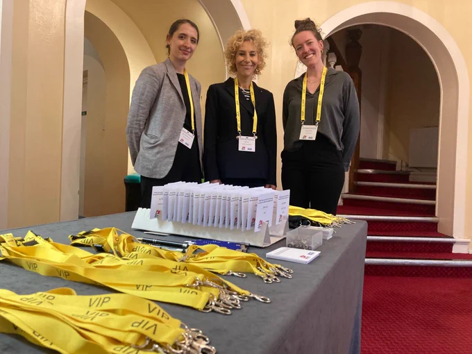 Three event staff wearing yellow lanyards stand behind a table filled with VIP badges and registration materials in a venue with arched doorways.
