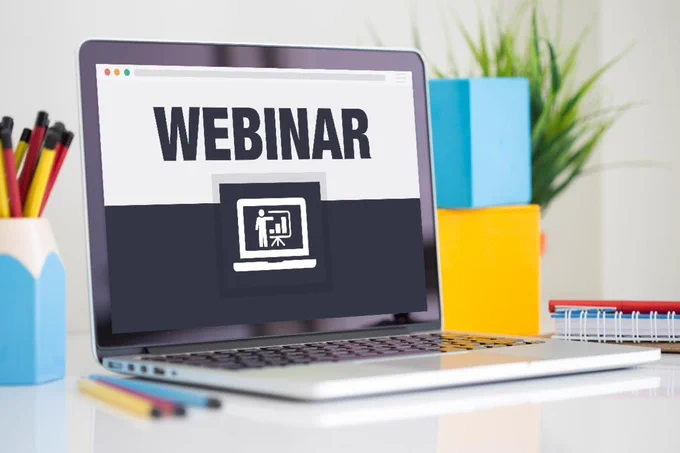 Silver laptop displaying "WEBINAR" on its screen sits on a white desk next to a pencil holder and colorful office supplies with a green plant in the background.