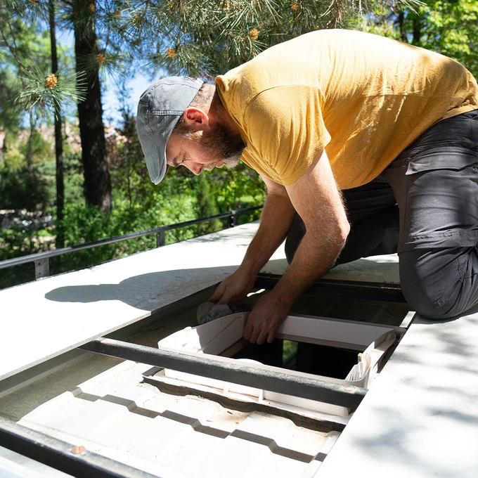 A person in a yellow shirt and cap is installing or repairing a skylight on the roof of a vehicle. They are outdoors, surrounded by trees.