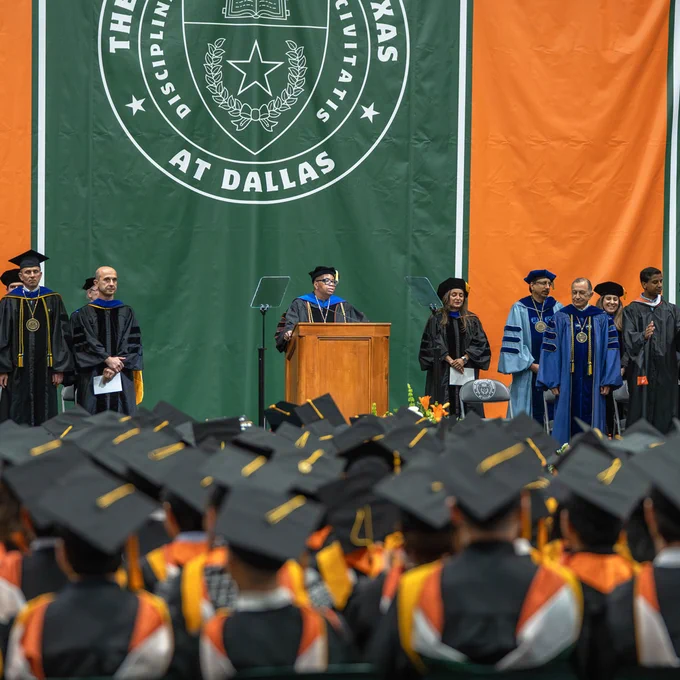 Dean Adams stands at a podium on a stage, addressing a graduation ceremony at The University of Texas at Dallas. Several individuals in academic attire are standing on stage. Numerous graduates in caps and gowns are seated in the foreground.